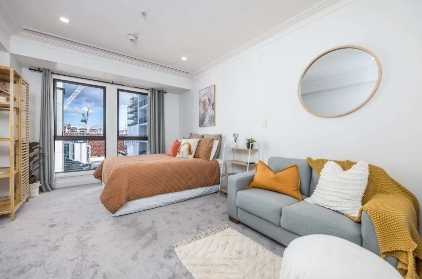 CBD Studio Getaway: Bright studio with a queen bed in a rust-orange cover, a grey sofa with a mustard throw, a round timber mirror and tall windows looking out over the city